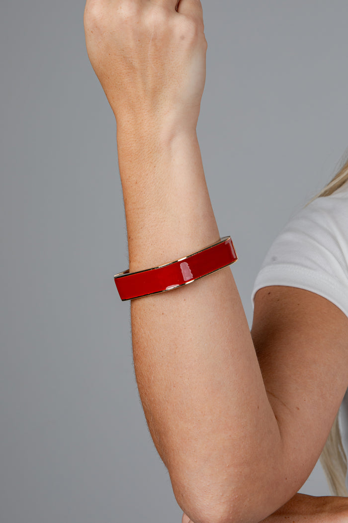 A close-up of a person's forearm wearing a glossy red rectangular cuff bracelet with a gold-toned metal edge, against a plain gray background.