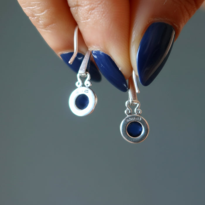 Silver earrings with blue stones held by a hand with blue nail polish on a gray background