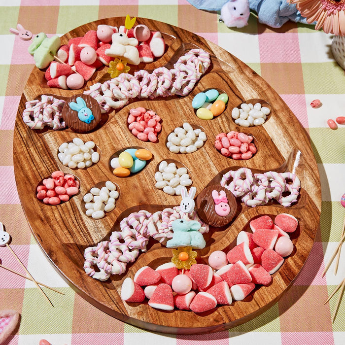 A charcuterie serving board designed to look like an Easter egg, with various candy-like items embedded in it, displayed on a table with a checkered cloth.