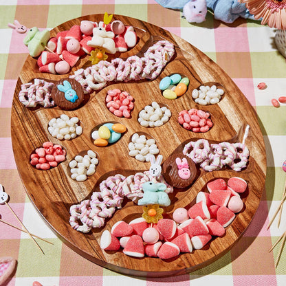 A charcuterie serving board designed to look like an Easter egg, with various candy-like items embedded in it, displayed on a table with a checkered cloth.