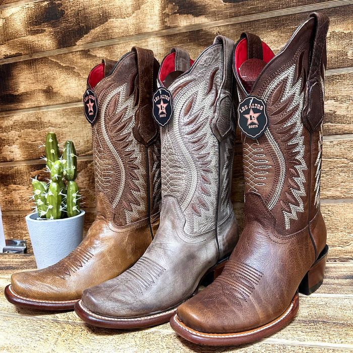 Three pairs of Los Altos Women’s Dubai Western Boots in varying shades of brown displayed on a wooden background with a cactus beside them.