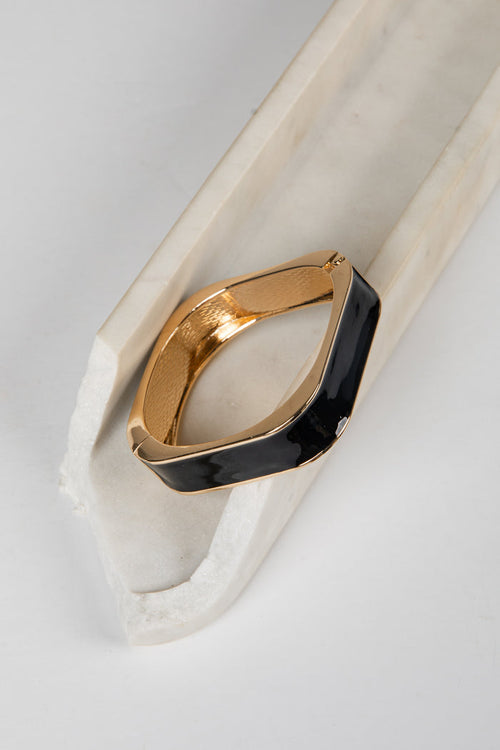 A gold-toned metal cuff bracelet with a wide, curved black enamel inlay, displayed on a white marble prop against a light gray background.