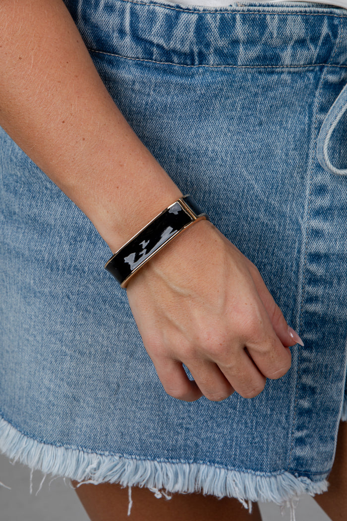 A close-up of a person's hand wearing a black rectangular cuff bracelet with a silver-toned metal frame and white abstract line details, paired with a light blue denim skirt.