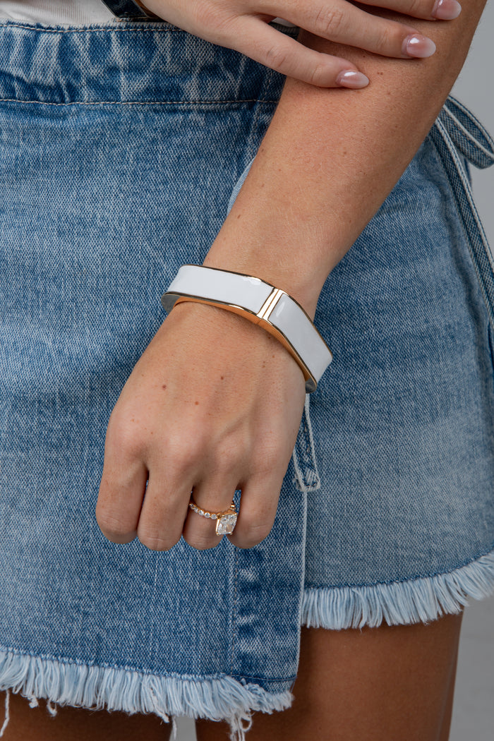 A close-up of a person wearing a wide white cuff bracelet with a gold-toned metal band and a gold accent bar, paired with a denim skirt.